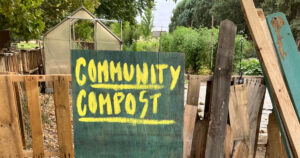 Green sign with yellow lettering “Community Compost” in front of a wood fence, greenhouse, and lush greenery.
