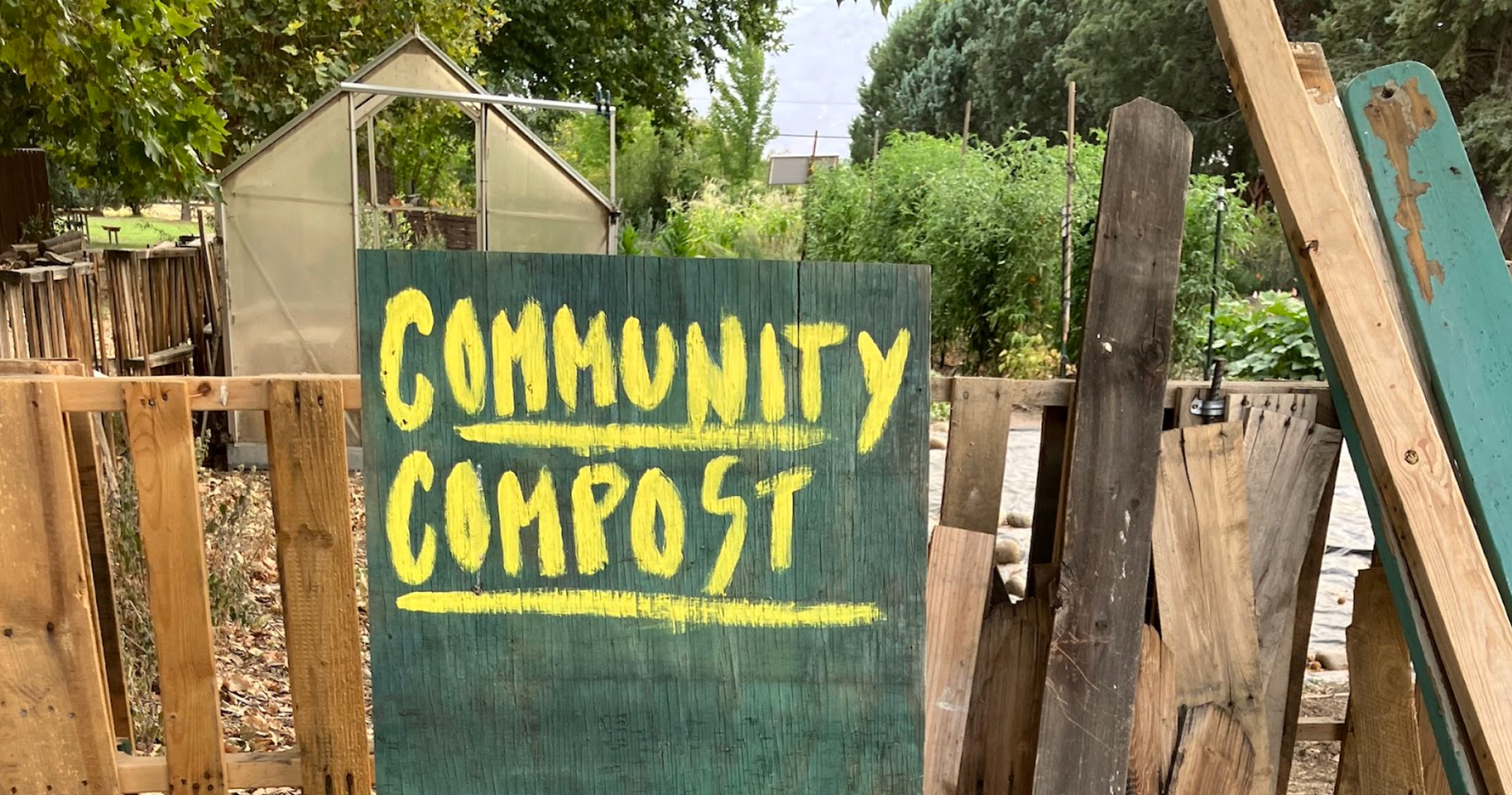 Green sign with yellow lettering “Community Compost” in front of a wood fence, greenhouse, and lush greenery.