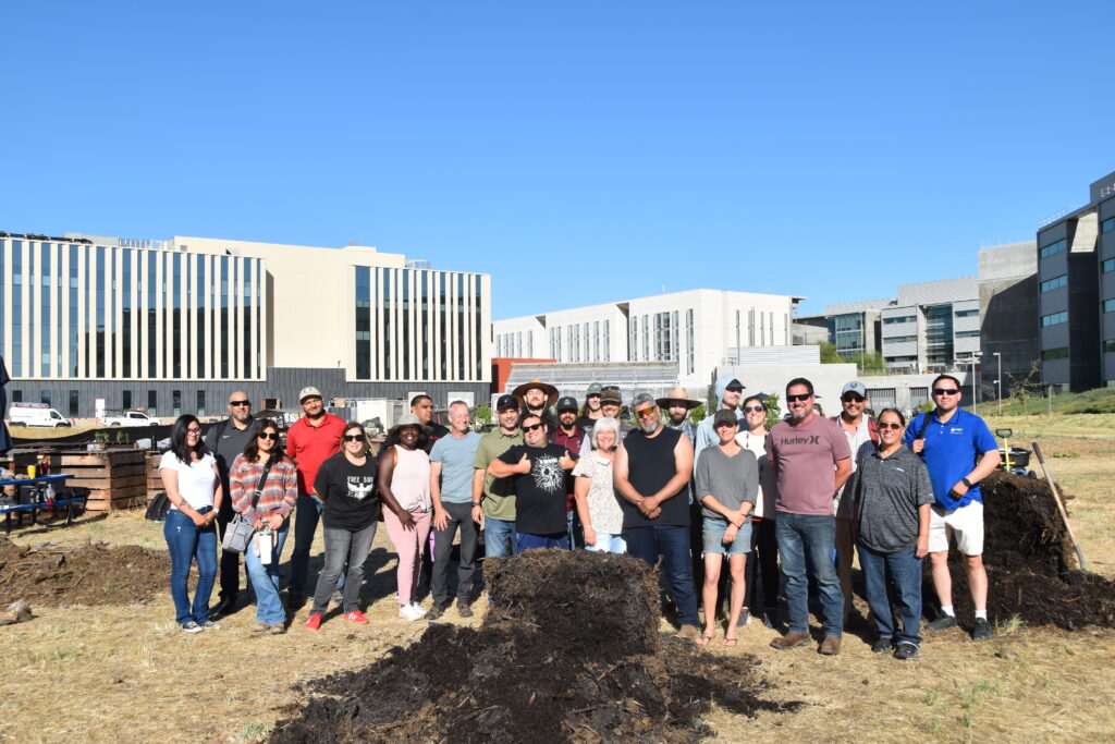Twenty four people standing in a field with compost piles.